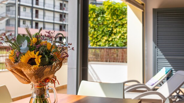 A room with a table, a vase of flowers, and a view of buildings and greenery outside.