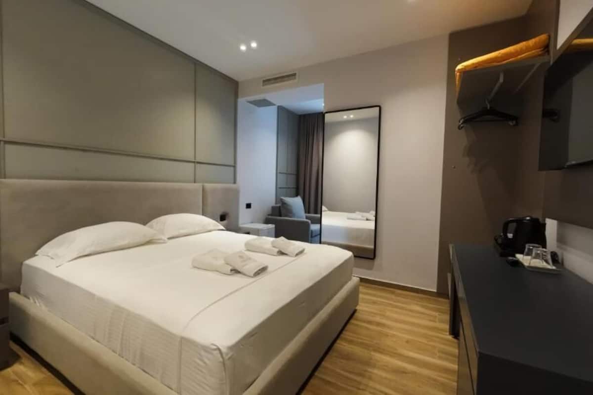 A modern hotel room with a large bed, a bedside table, a desk with a chair, a mirror, and a wardrobe.