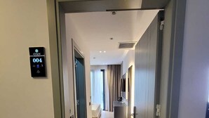 En hotellkorridor med en digital kodpanel som visar rum nummer 004, en säng, ett litet pentry och en spegel på väggen.