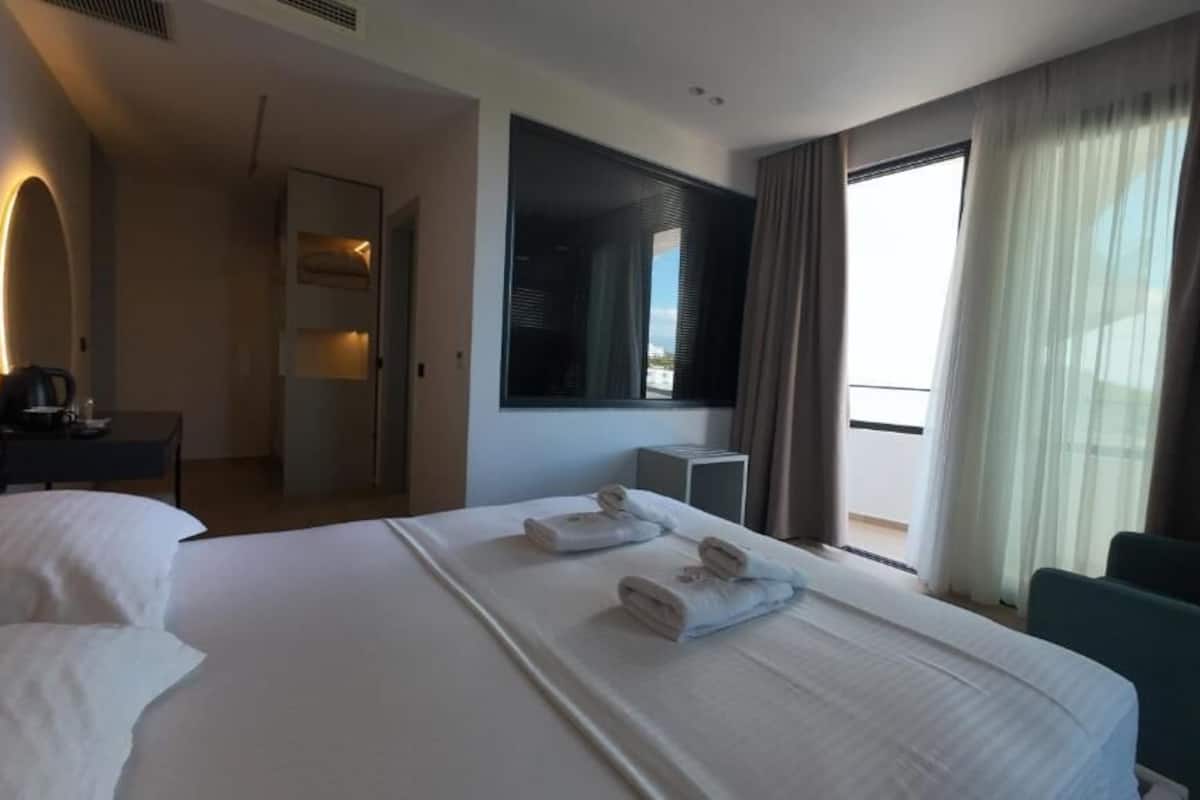 A hotel room with a large bed, a desk, a chair, a TV, and a balcony with curtains.