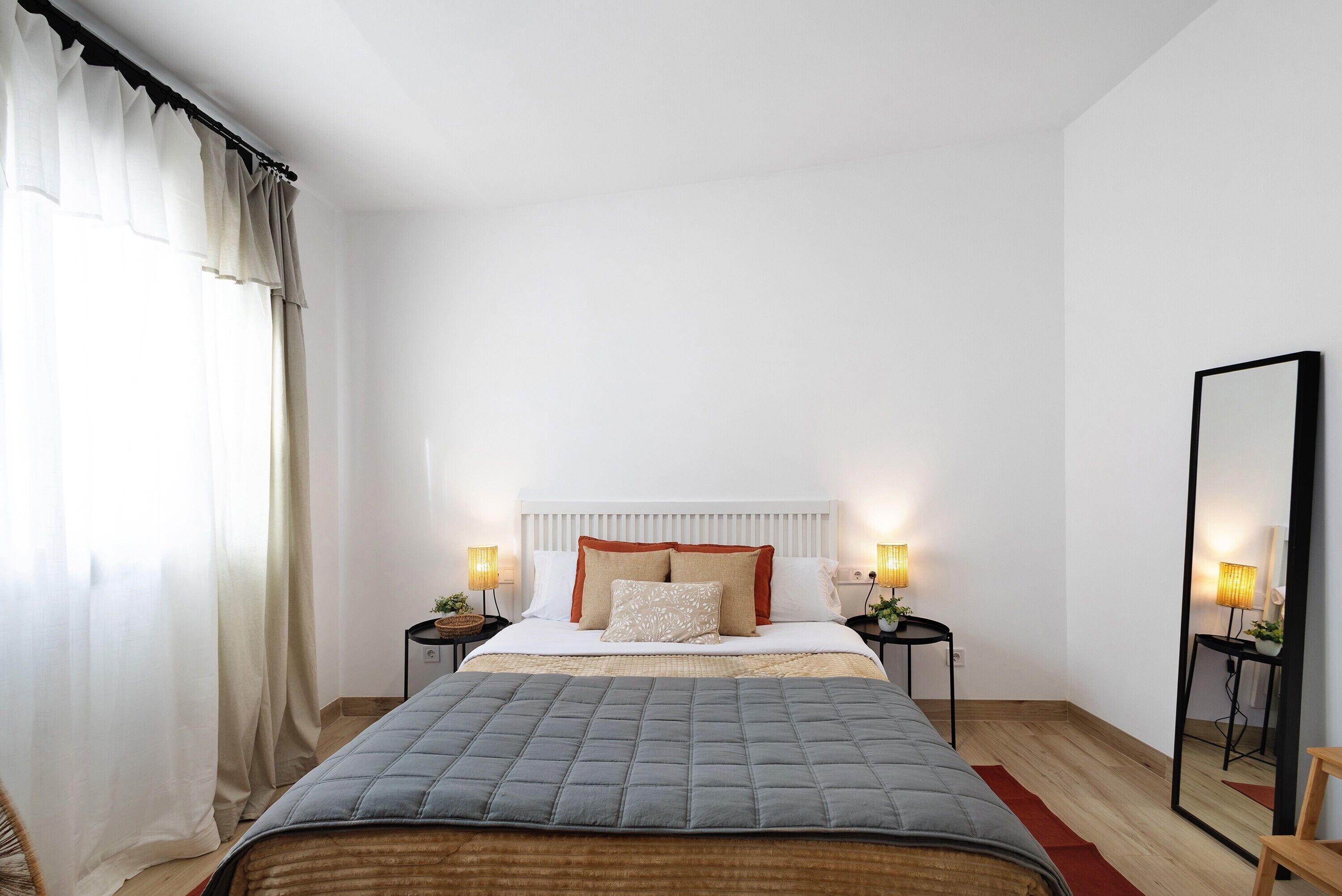 Un dormitorio con una cama, dos mesitas de noche con lámparas, un espejo y una ventana con cortinas.