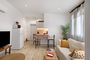 Ein modernes Wohnzimmer mit einem Flachbildfernseher, einer Couch mit gemusterten Kissen, einem Essbereich mit einem Tisch und einer Küche im Hintergrund.