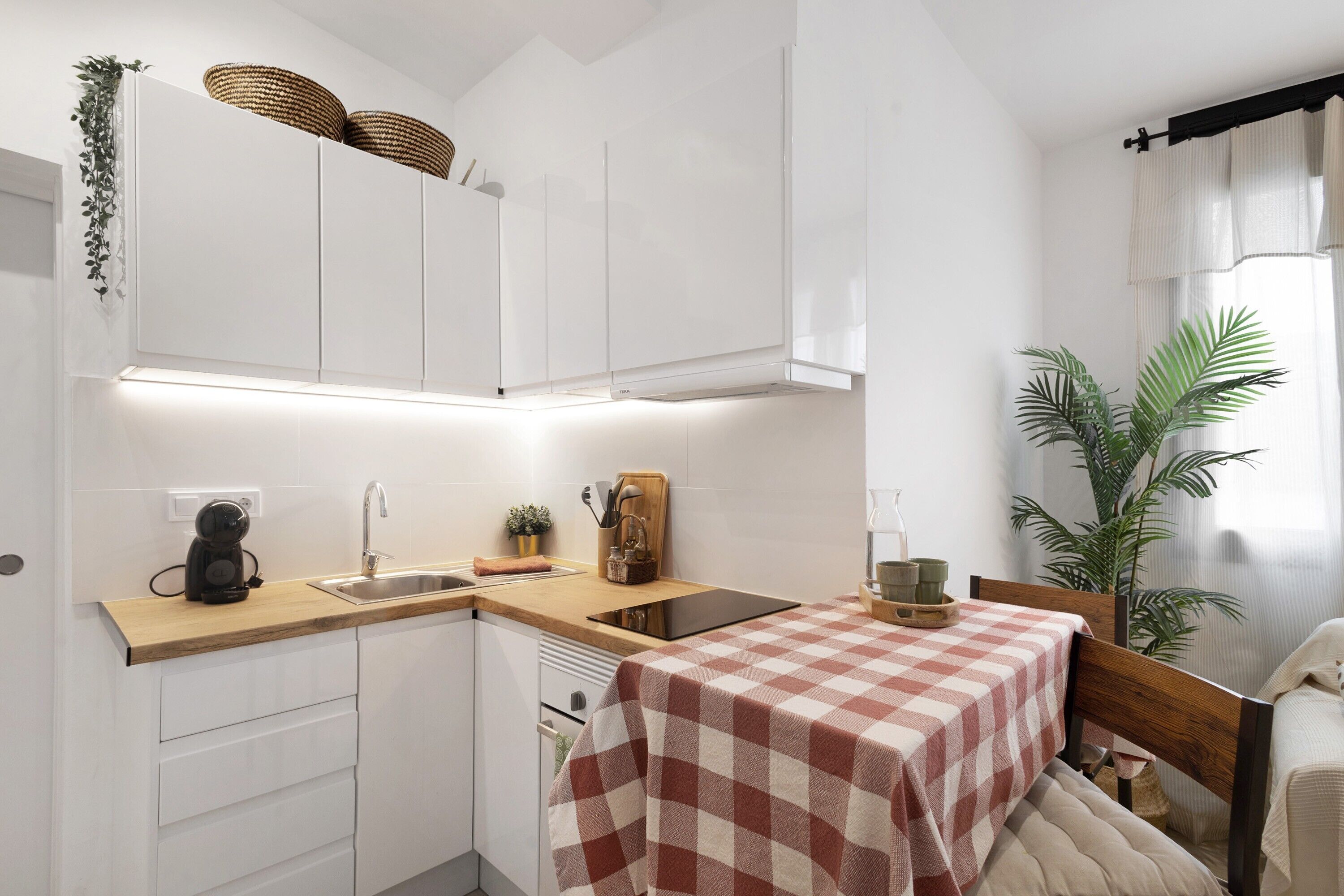 Una cucina compatta con mobili bianchi, un piano di lavoro in legno, una tovaglia a quadri e una pianta in vaso.