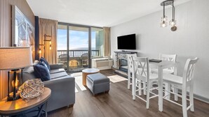 A modern living room with a sofa, ottoman, and dining table with chairs. There is a fireplace, a television, and a large window with a view. - Sun & Sand Waiting for You: Oceanfront Ste 4th Flr (Myrtle Beach)