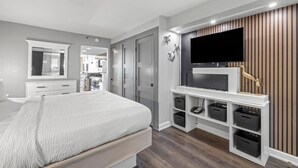 A modern bedroom with a large bed, a TV mounted on a wooden panel wall, and a white console table with storage baskets. - Sun & Sand Waiting for You: Oceanfront Ste 4th Flr (Myrtle Beach)