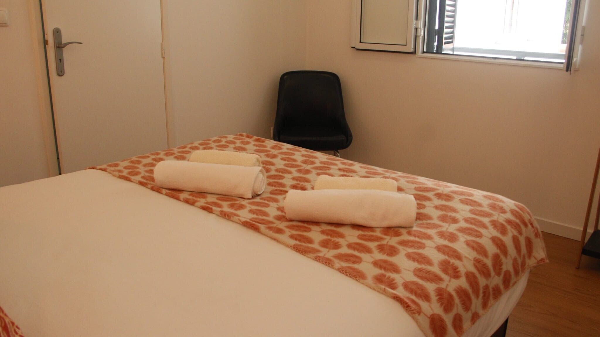 Une chambre d’hôtel avec un lit, deux serviettes enroulées, une chaise noire, une fenêtre et une porte.