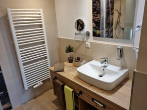Un baño con un lavabo blanco, un espejo, un perchero para toallas, una pequeña planta en maceta y una ducha con vistas a una cascada.