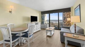 Hotelkamer met een eettafel, stoelen, een bank, een televisie, een klein bijzettafeltje en een balkon met uitzicht.