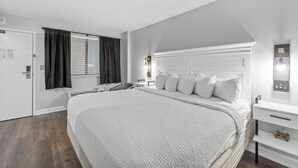 A modern hotel room with a large bed, a bedside table, a nightstand, a window with curtains, and a wall-mounted control panel.