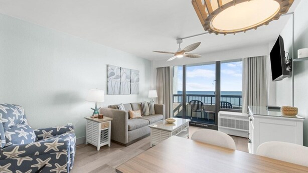 A modern living room with a sofa, a dining table, and a ceiling fan. - Seaside Tranquility Spacious Suite 5th Floor (Myrtle Beach)