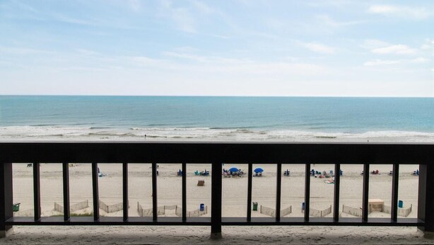 Vista a la playa desde un balcón con barandilla, playa de arena y océano.