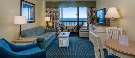 A hotel room with a sofa, a small table, a television, and a view of the ocean.