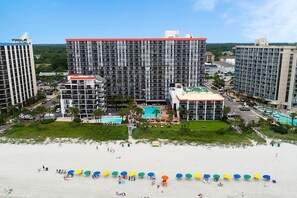 A hotel with multiple high-rise buildings, a central pool area, and a beach with umbrellas.