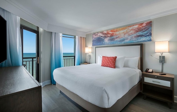 A hotel room with a large bed, a nightstand, a TV, and a view of the ocean.