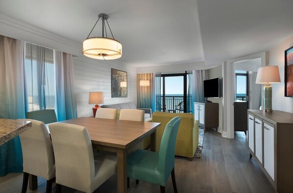 A hotel room with a wooden dining table, beige and teal chairs, a sofa, a TV, and a minibar. - Elegance Oceanfront 3 BR Condo 12th Floor (Myrtle Beach)