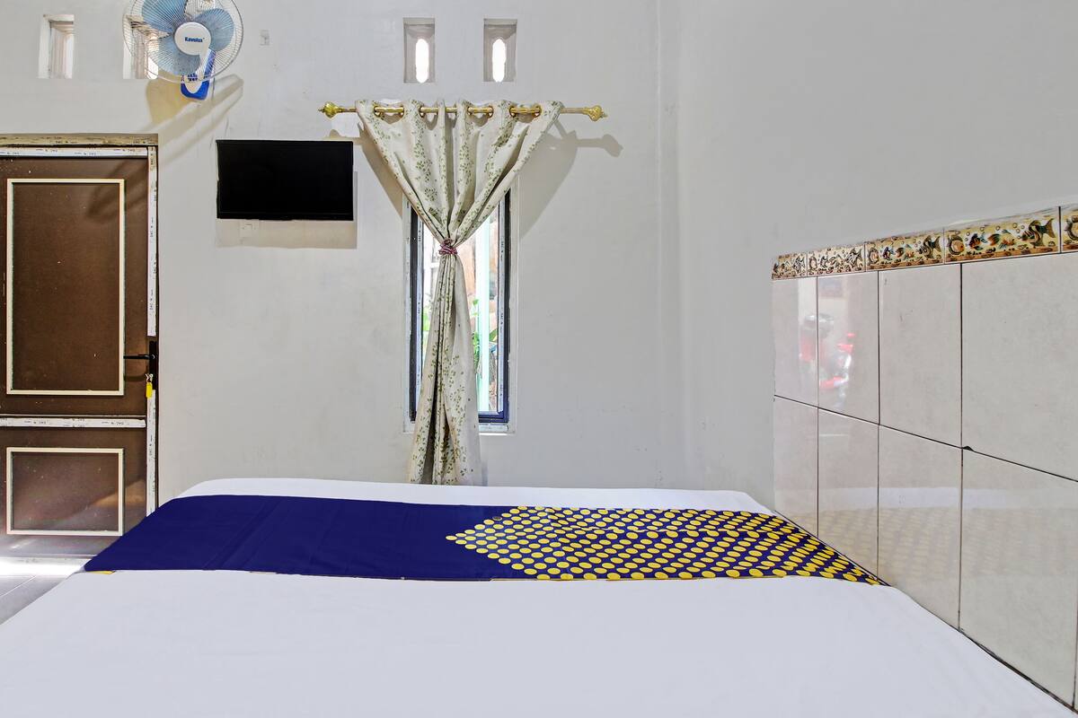 Une chambre d’hôtel avec un lit, une télévision, une fenêtre avec des rideaux et un ventilateur fixé au mur.
