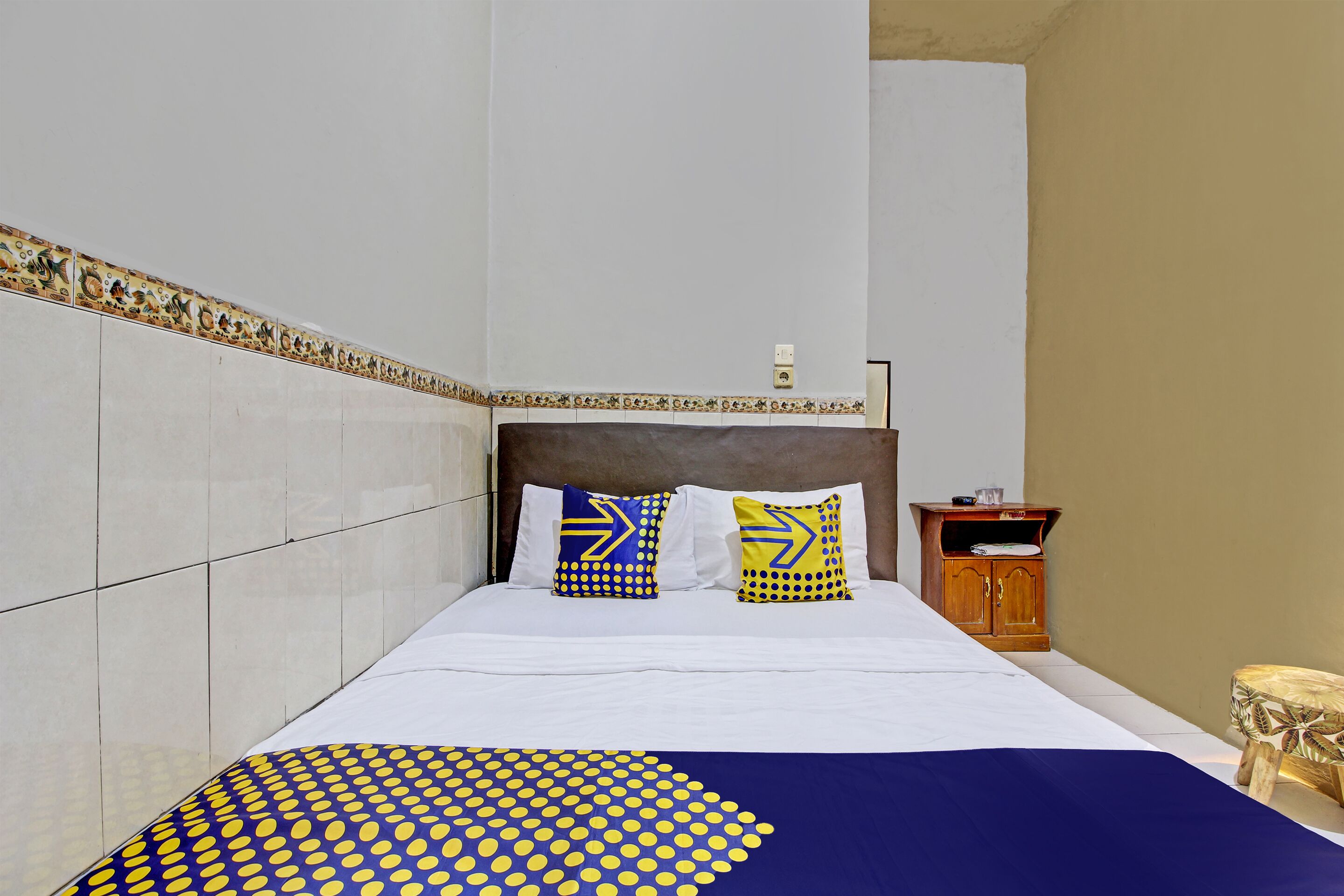 A neatly made bed with white linens and two pillows, a wooden nightstand with a lamp, and a small wooden cabinet.