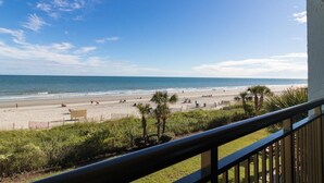 Uitzicht op een strand vanaf een balkon met een hekwerk, palmbomen en een heldere lucht.