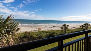 A beach view from a balcony with a railing, palm trees, and a clear sky.