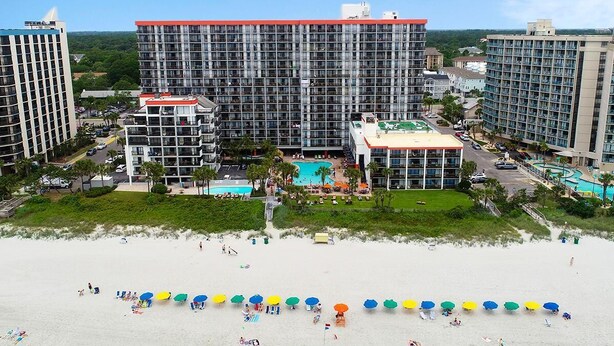 Un hotel con varios edificios altos, un área de piscina central y una playa con sombrillas.