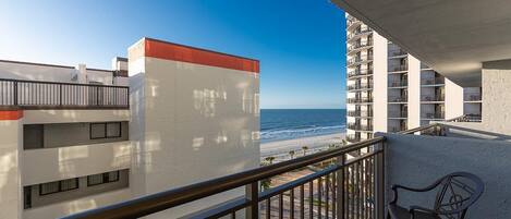 A balcony with a view of the beach and ocean, a chair, and a building with balconies.