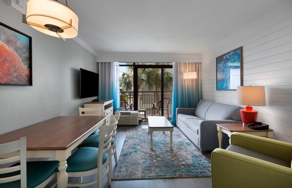 A modern hotel room with a dining area, a sofa, a TV, and a balcony with a view of palm trees. - Partial Ocean View Suite on the 8th Floor (Myrtle Beach)