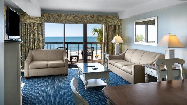 Camera d'hotel con una grande finestra vista mare, un divano, una sedia, un tavolino e una TV.