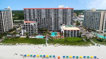 A hotel with multiple high-rise buildings, a central pool area, and a beach with umbrellas.