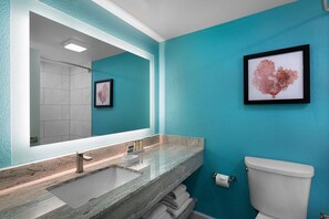 Une salle de bain avec un mur turquoise, un grand miroir, un lavabo avec un plan de toilette en marbre, des toilettes et une œuvre d’art encadrée.