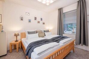 Una cama bien hecha con estructura de madera, una mesita de noche con lámpara y una ventana con cortinas.