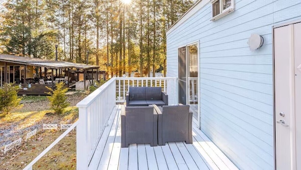 A covered outdoor seating area with a deck, a light blue building, and a forested backdrop.