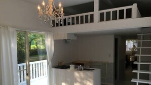 A spacious living area with a white sofa, a dining table, and a chandelier. There is a balcony with a white railing, and a staircase leading to an upper level.