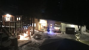 A snow-covered outdoor area with a building featuring a wooden deck and railings, illuminated by string lights.