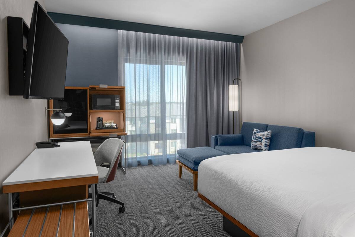 A hotel room with a bed, a desk, a chair, a TV, a bookshelf, and a window with curtains.