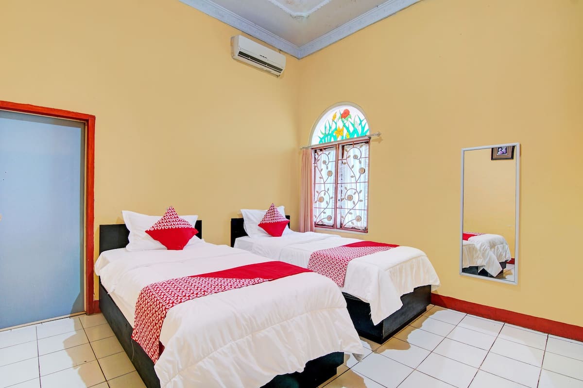 A hotel room with two beds, a window with a stained glass design, a mirror, and an air conditioning unit.