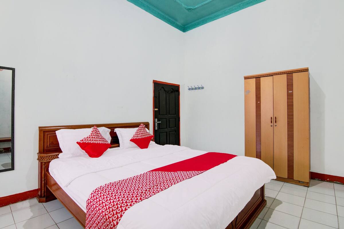 A neatly made bed with a red and white patterned blanket, a wooden wardrobe, and a black door in a room with white walls and a teal ceiling.