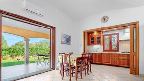 A dining area with a table and chairs, a kitchen with wooden cabinets, and a view of greenery outside.