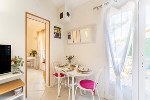 A dining area with a table set for four, white chairs, a television on a shelf, and a door leading to a garden.