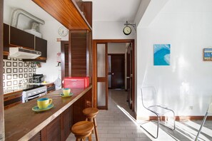 A kitchen with wooden cabinets, a stove, and a red appliance on the counter. There are two stools and a clear chair. A clock and a framed picture are on the wall.