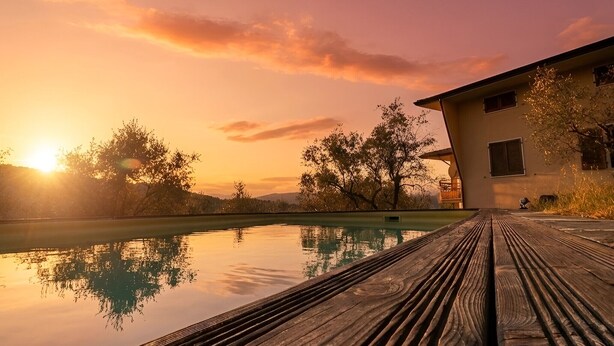 Une terrasse en bois qui mène à une piscine, avec un coucher de soleil en arrière-plan.