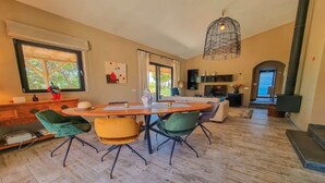 Amplia sala de estar con una gran mesa de comedor de madera, sillas verdes y amarillas, chimenea y un ventanal con vistas a árboles.