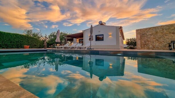 Una piscina con agua cristalina que refleja el cielo, un edificio blanco con una zona techada y un muro de piedra.
