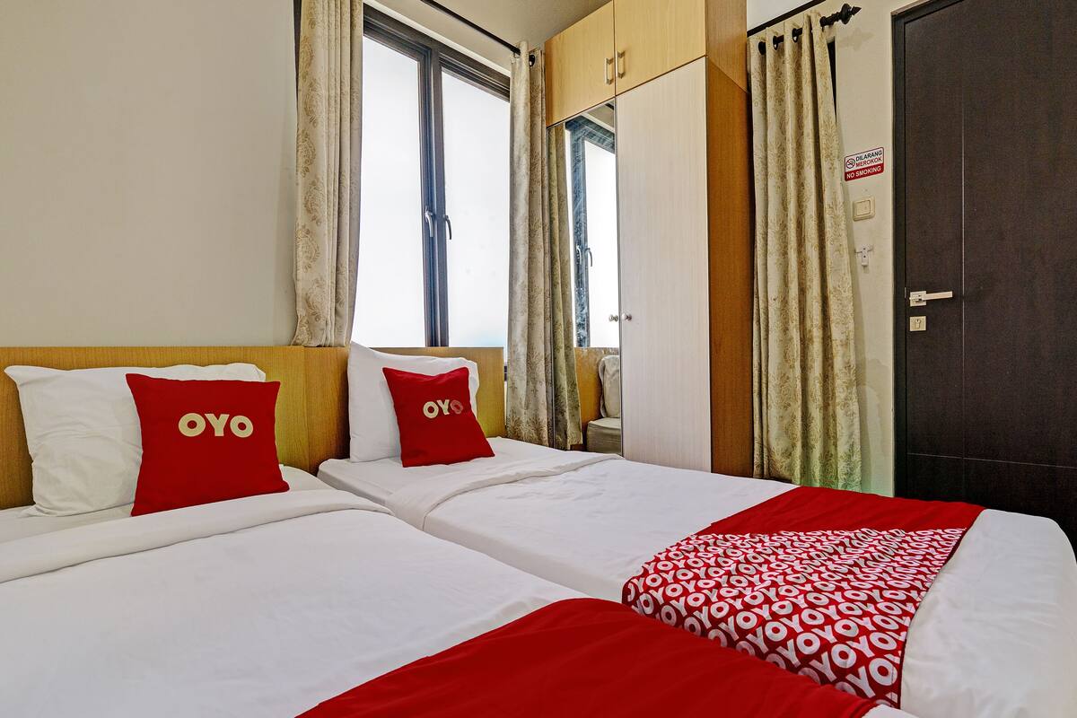 Habitación de hotel con dos camas, cada una con ropa blanca y almohadas rojas. Amplia ventana con cortinas, armario y puerta con letrero de 'No Fumar'.