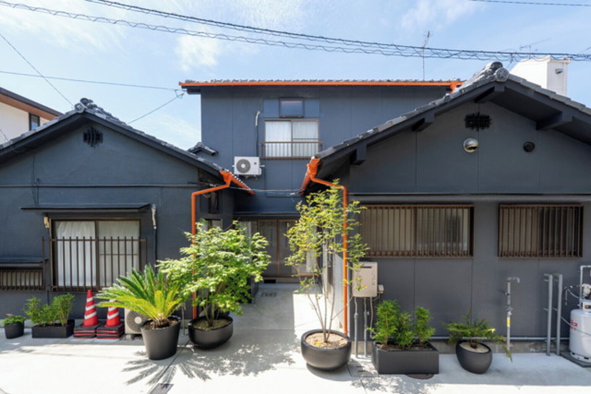 A residential building with a grey exterior, two visible windows, and a small air conditioning unit. There are potted plants in front of the building.