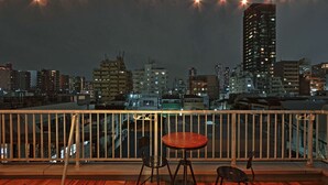 A rooftop terrace with a wooden floor, metal railing, and a table with two chairs. The terrace overlooks a cityscape with various buildings and a tall skyscraper.