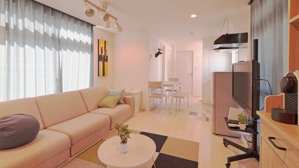 A modern living room with a sofa, coffee table, and a TV. There is a kitchen area with a refrigerator and a microwave. The room has a ceiling light fixture and a door leading to another room.