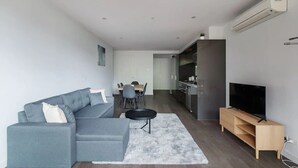 A modern living room with a grey sectional sofa, a small round table, a flat-screen TV on a wooden stand, and a kitchen area in the background.