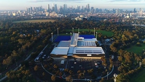 An aerial view of a modern building with a unique roof design, surrounded by a parking lot and greenery, with a city skyline in the background.