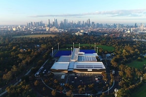 Une vue aérienne d’un bâtiment moderne doté d’un toit au design original, entouré d’un parking et de verdure, avec une vue sur les gratte-ciel de la ville en arrière-plan.