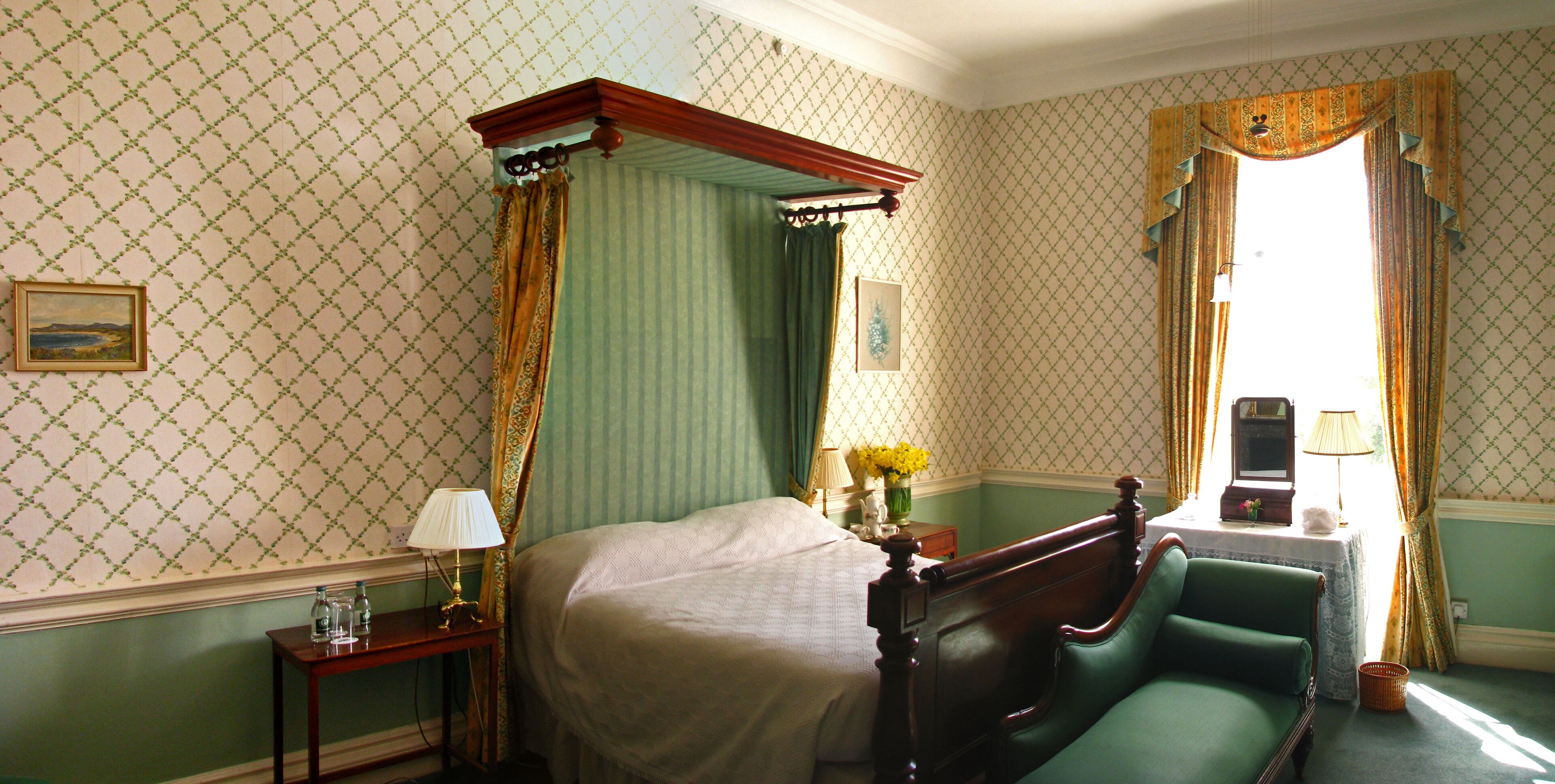 Une chambre avec un lit à baldaquin, un fauteuil vert, une table de chevet avec une lampe et une fenêtre avec des rideaux.